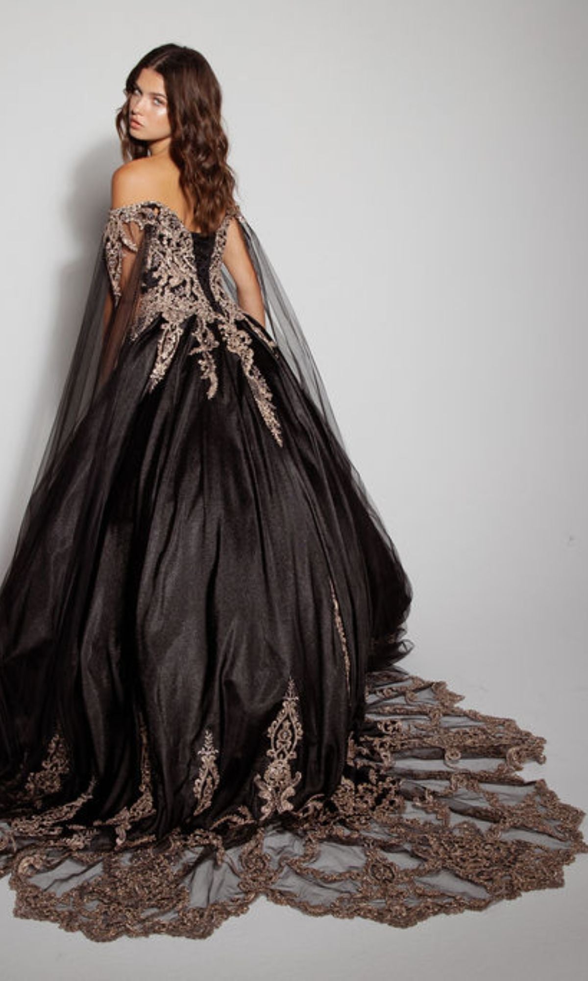 A woman models the Quinceanera Dress EK1002 By Eureka—an off-the-shoulder black gown with gold embroidery, lace-up back, and a long train—standing against a plain background.